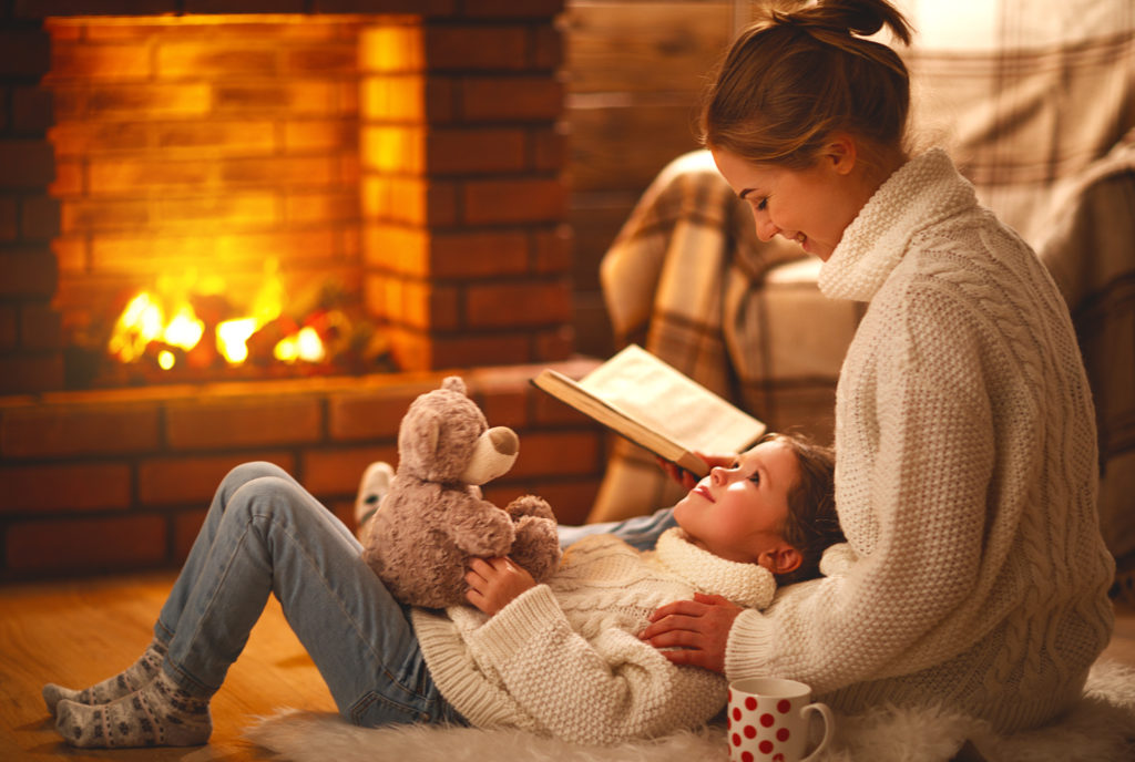 family mother and child daughter reading book on winter evening by fireplace